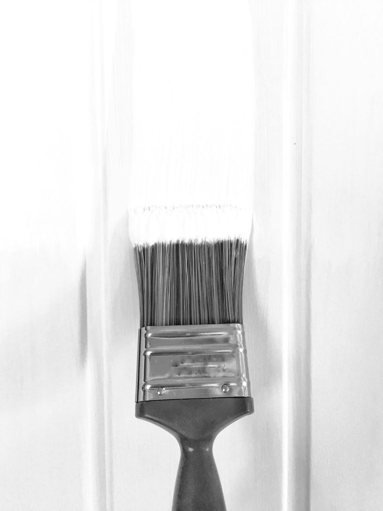 A close up of a paint brush painting and decorating a white wooden door with white paint or emulsion